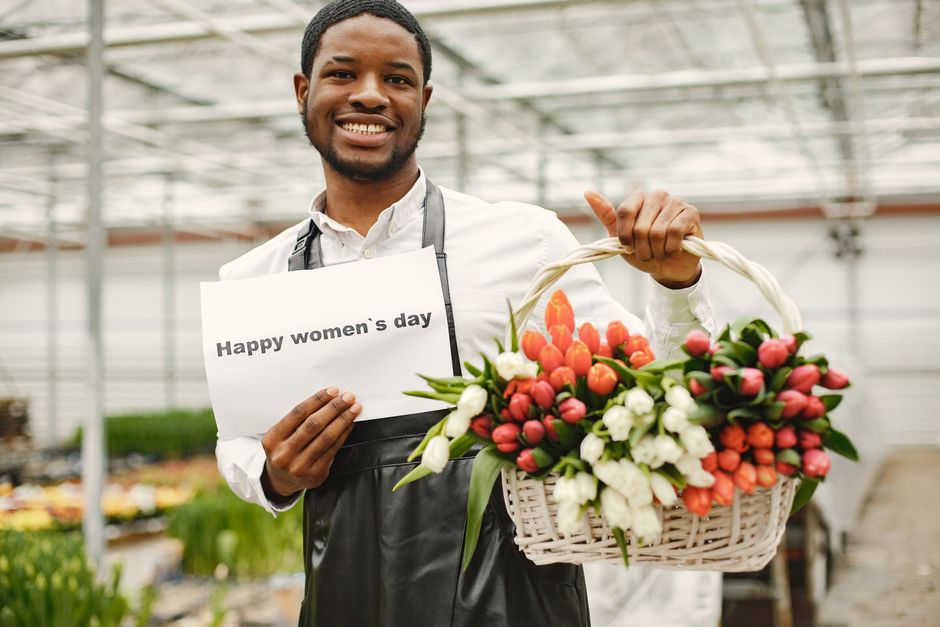 A smiling male florist with dark curly hair and a beard, wearing a white shirt and a black apron, stands inside a greenhouse holding a festive basket filled with fresh pink tulips and white freesias.
